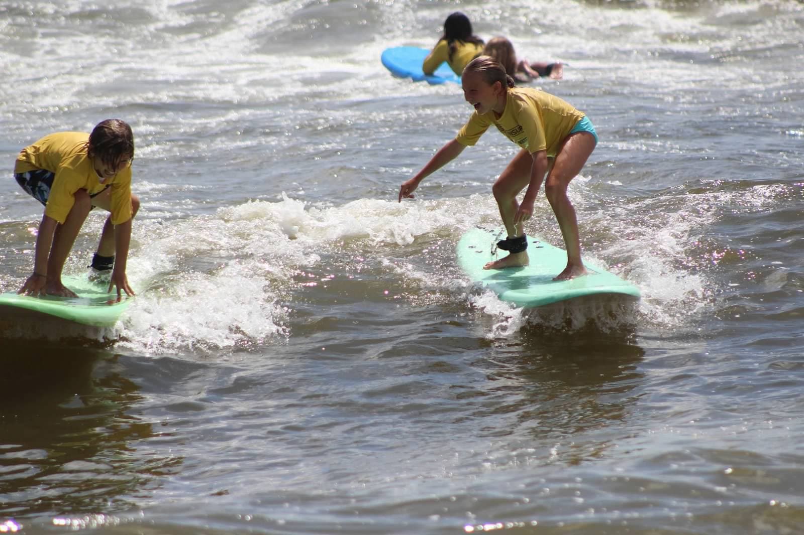 Two day camp children in yellow rash guards riding foam boards in the surf at The Sands